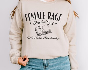 May include: A beige sweatshirt featuring the text "FEMALE RAGE Readers' Club" in black, and an illustration of an open book. The words "Worldwide Membership" are printed below the book. The sweatshirt is worn with blue jeans.