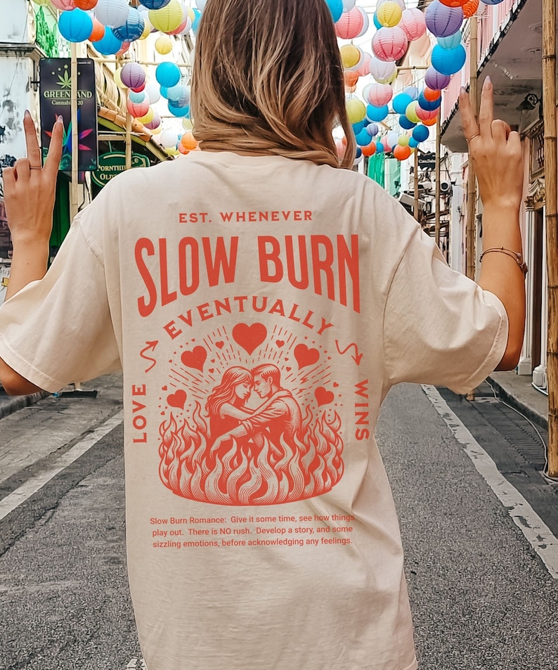 Op de afbeelding: Cr&egrave;mekleurig T-shirt met de rode tekst "EST. WHENEVER SLOW BURN EVENTUALLY LOVE WINS". De afbeelding toont een stel dat elkaar omhelst in vlammen, omringd door harten. Het shirt heeft ook de tekst "Slow Burn Romance" en een korte beschrijving.