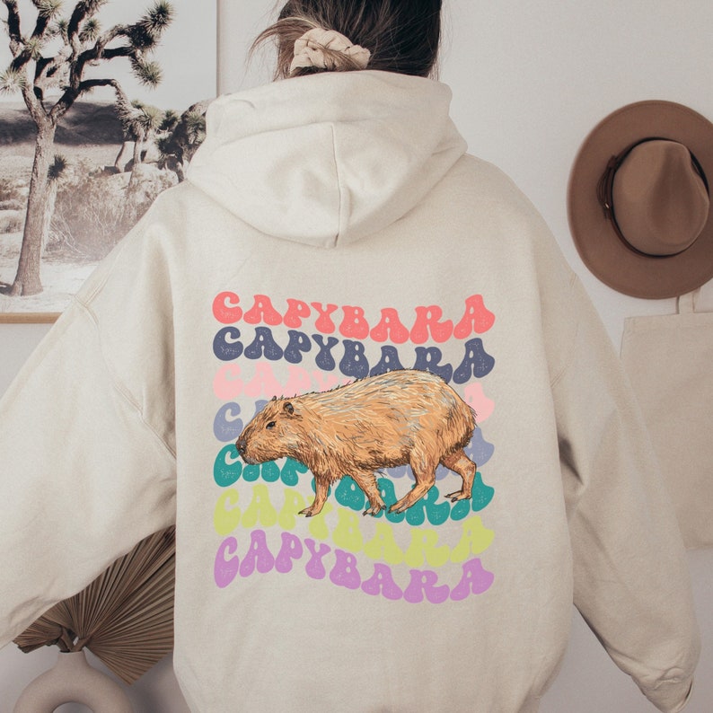 K&ouml;nnte beinhalten: Ein beiger Hoodie mit einem Grafikdruck eines Capybaras und dem Wort "Capybara", das in einem Regenbogen aus Farben wiederholt wird.