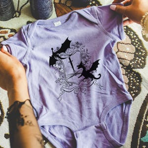 May include: A lavender baby onesie with a black and white graphic of a dragon and a sword on a shield. The graphic is surrounded by floral details.