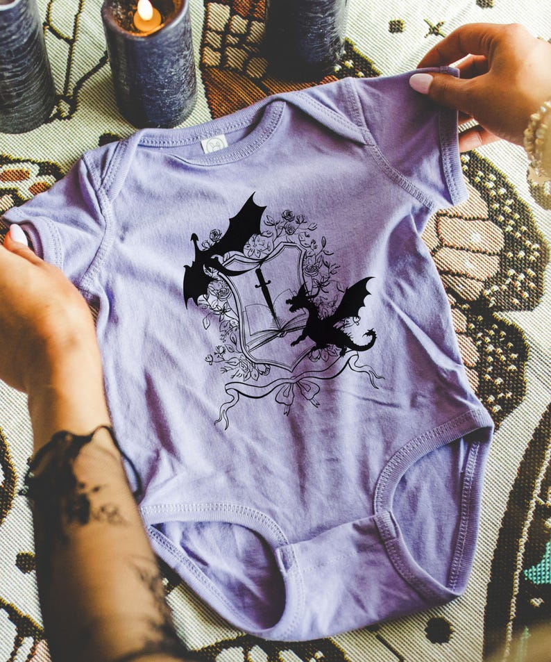May include: A lavender baby onesie with a black and white graphic of a dragon and a sword on a shield. The graphic is surrounded by floral details.