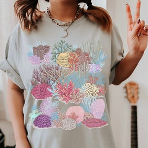 May include: A light blue t-shirt featuring a colourful illustration of coral reefs. The illustration depicts various types of coral, seaweed, and other marine life.