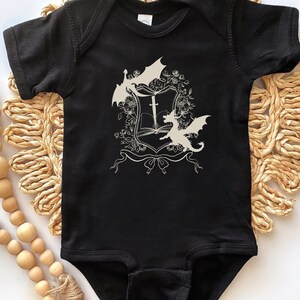 May include: Black baby onesie with a white design of a shield with a sword and two dragons.