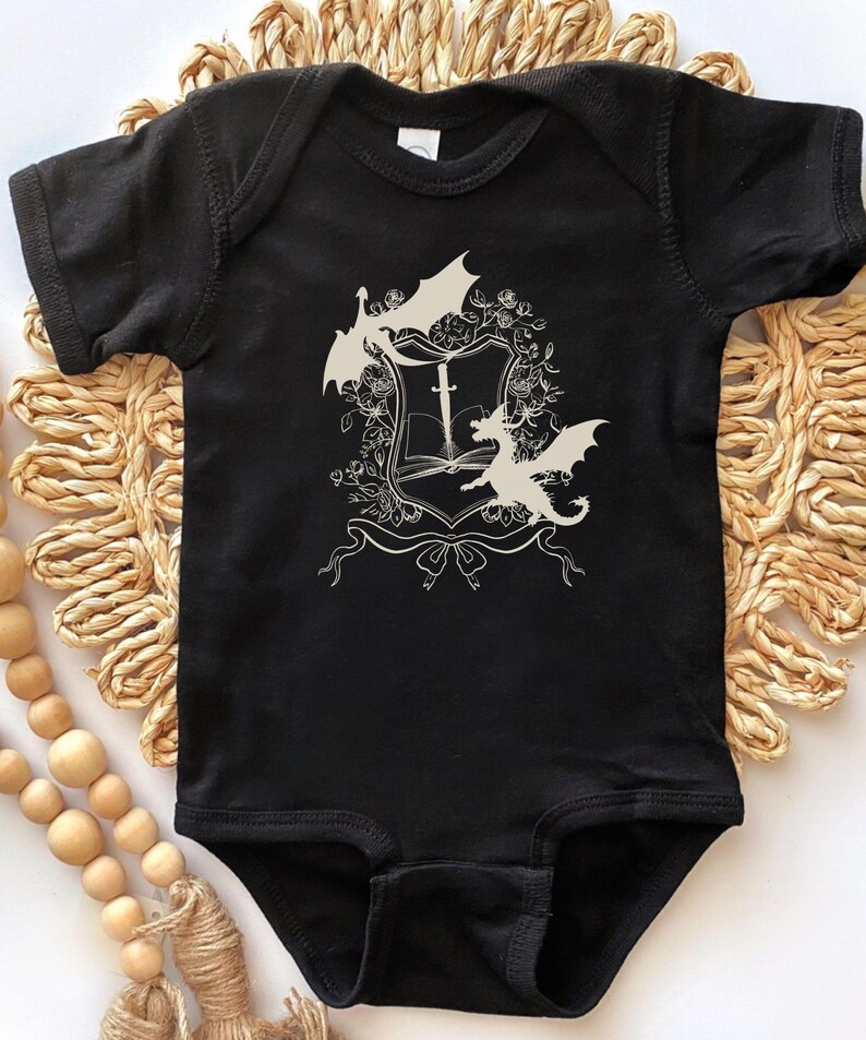 May include: Black baby onesie with a white design of a shield with a sword and two dragons.