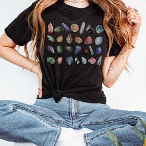 May include: A black t-shirt with a colorful print of various gemstones. The shirt is worn by a person sitting with their legs crossed, wearing blue jeans.