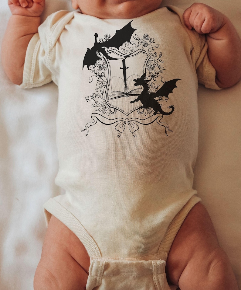 May include: A white baby onesie with a black and white graphic of a shield with a sword, book, and two dragons.
