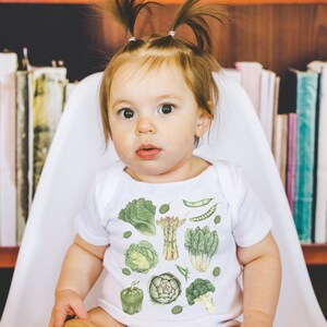 May include: A white baby onesie with a green and white print of various vegetables, including asparagus, artichokes, and leafy greens. The onesie is worn by a baby with brown hair in pigtails.