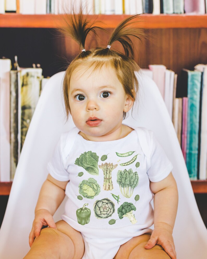 May include: A white baby onesie with a green and white print of various vegetables, including asparagus, artichokes, and leafy greens. The onesie is worn by a baby with brown hair in pigtails.