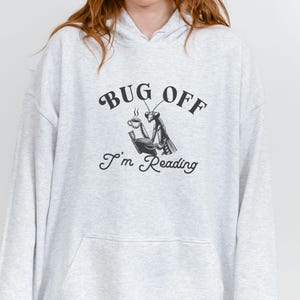 May include: Light gray hoodie with a front graphic. The graphic features a praying mantis reading a book and drinking coffee, with the text "BUG OFF I'm Reading" in black. The hoodie has a front pocket and a hood.