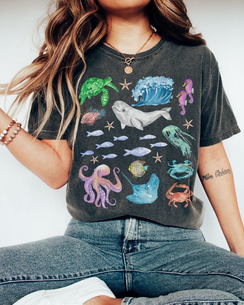 May include: A dark grey t-shirt with a colourful ocean-themed graphic print. The print features a variety of sea creatures, including a whale, octopus, sea turtle, jellyfish, and fish. The text "Mer Oc&eacute;an" is printed on the right sleeve.