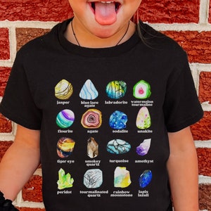 May include: Black t-shirt featuring a colorful graphic of various gemstones, including jasper, amethyst, and turquoise. Each stone is labeled with its name. The shirt is worn by a child.