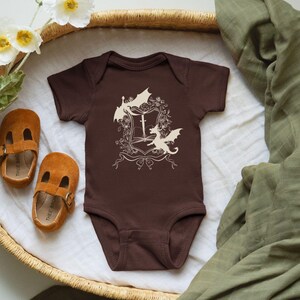May include: A brown baby onesie with a white design of a book with a sword and three dragons. The onesie is in a wicker basket with a green blanket.