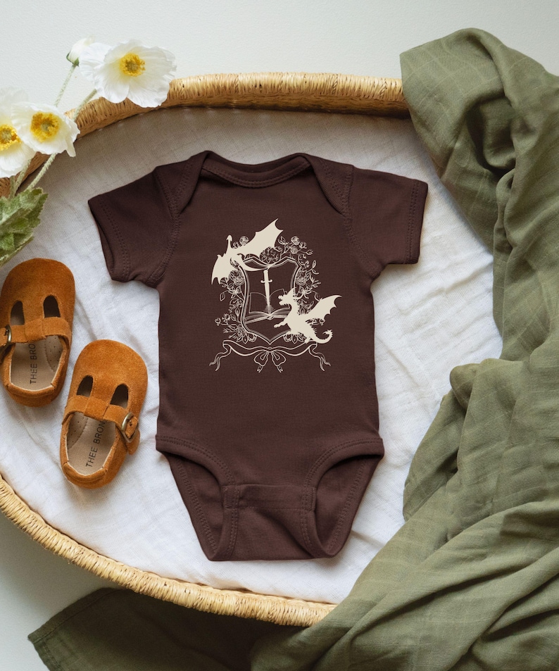 May include: A brown baby onesie with a white design of a book with a sword and three dragons. The onesie is in a wicker basket with a green blanket.
