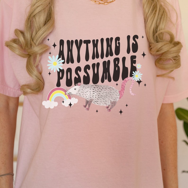 May include: Pink t-shirt with the text "ANYTHING IS POSSUMBLE" in a retro font. A watercolor possum, rainbow, and daisies are featured in the design. Black star accents complete the graphic.