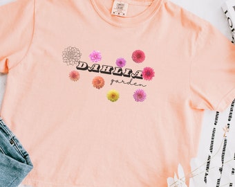 May include: A peach-coloured t-shirt with a graphic design of colourful dahlias and the text "DAHLIA garden" in a retro font.