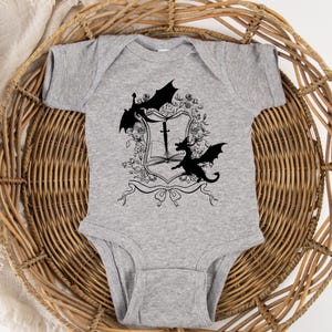 May include: A gray baby onesie with a black and white graphic of two dragons flanking a shield with a sword and a book. The shield is surrounded by floral designs.
