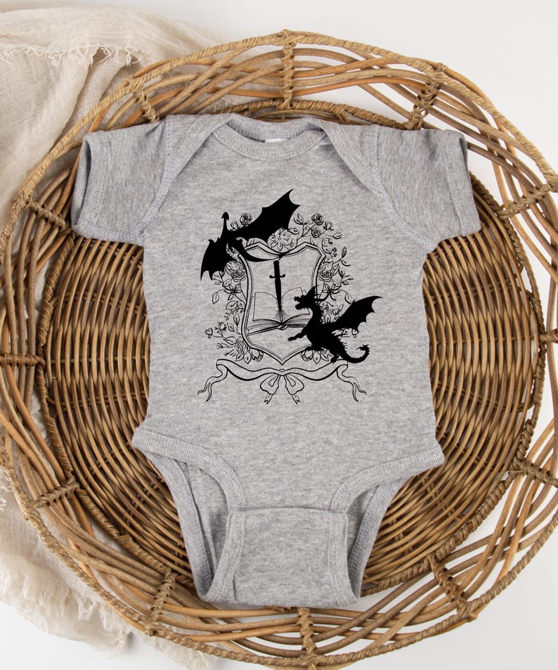 May include: A gray baby onesie with a black and white graphic of two dragons flanking a shield with a sword and a book. The shield is surrounded by floral designs.