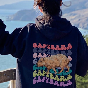 K&ouml;nnte beinhalten: Ein schwarzer Hoodie mit einem Aufdruck eines Capybaras und dem Wort "Capybara", das in einem Regenbogen-Farbmuster wiederholt wird.