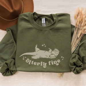 Puede incluir: Una sudadera verde de manga larga con un gr&aacute;fico blanco de una nutria tumbada de espaldas con estrellas a su alrededor. El texto "otter-ly fine" est&aacute; debajo de la nutria.