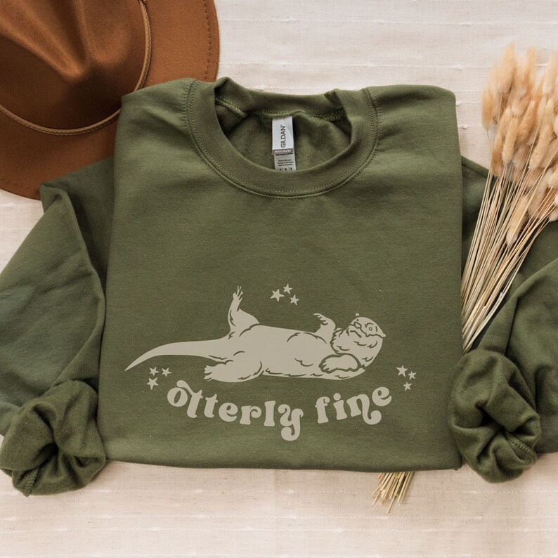 Puede incluir: Una sudadera verde de manga larga con un gr&aacute;fico blanco de una nutria tumbada de espaldas con estrellas a su alrededor. El texto "otter-ly fine" est&aacute; debajo de la nutria.