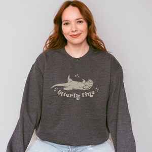 Puede incluir: Una sudadera gris oscuro con un gr&aacute;fico de una nutria acostada de espaldas con el texto "otter-ly fine" impreso debajo.