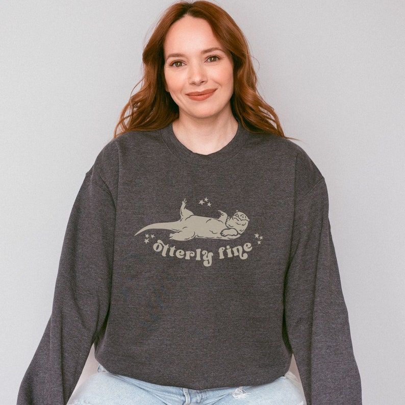 Puede incluir: Una sudadera gris oscuro con un gr&aacute;fico de una nutria acostada de espaldas con el texto "otter-ly fine" impreso debajo.