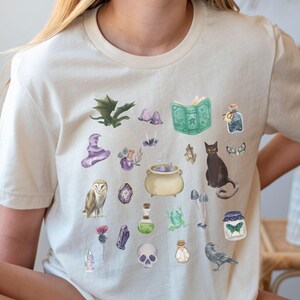 May include: Cream-colored t-shirt featuring watercolor illustrations of magical items. The design includes a witch's hat, potion bottles, a cauldron, a black cat, an owl, a skull, and a spell book. The overall theme is fantasy and witchcraft.
