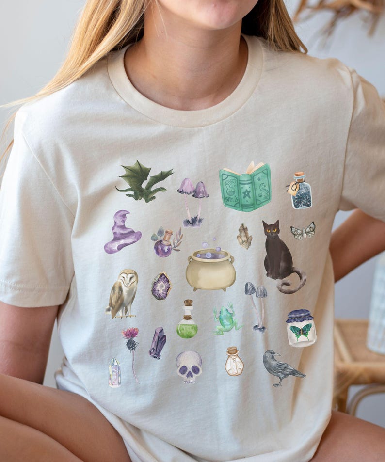May include: Cream-colored t-shirt featuring watercolor illustrations of magical items. The design includes a witch's hat, potion bottles, a cauldron, a black cat, an owl, a skull, and a spell book. The overall theme is fantasy and witchcraft.