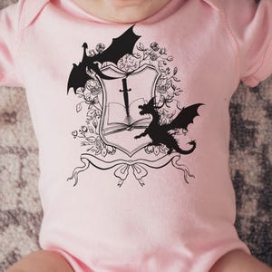 May include: Pink baby onesie with a black and white graphic of a book, sword, and two dragons. The graphic is surrounded by floral designs.