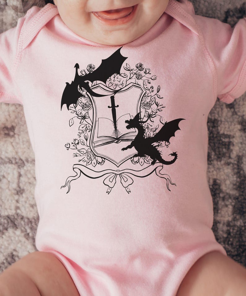 May include: Pink baby onesie with a black and white graphic of a book, sword, and two dragons. The graphic is surrounded by floral designs.