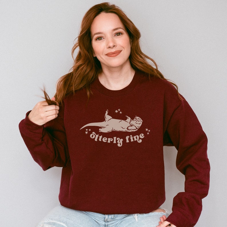 Puede incluir: Una sudadera color burdeos con un gr&aacute;fico de una nutria tumbada de espaldas con el texto "otter-ly fine" impreso debajo.