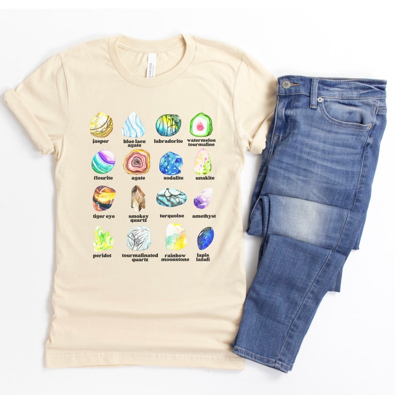 Op de afbeelding: Een beige T-shirt met een afbeelding van verschillende soorten edelstenen, waaronder jaspis, blauwe kant-agaten, labradoriet, watermeloen-tourmaline, fluoriet, agaat, sodaliet, unakiet, tijgeroog, rookkwarts, turquoise, amethist, peridoot, tourmalijnkwarts, regenboog-maansteen en lapis lazuli.