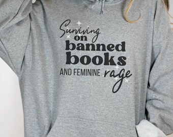 May include: Grey hooded sweatshirt with the text "Surviving on banned books and feminine rage" printed on the front.