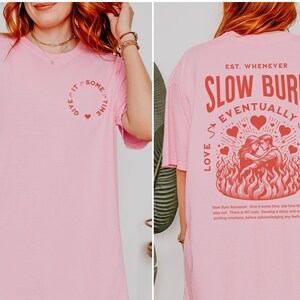 Op de afbeelding: Zachtroze T-shirt met rode tekst en afbeeldingen. De voorkant heeft een cirkelvormig ontwerp met de tekst "Give it some time" en een hart. De achterkant toont "Slow Burn Eventually Love Wins" met een stel in vlammen.