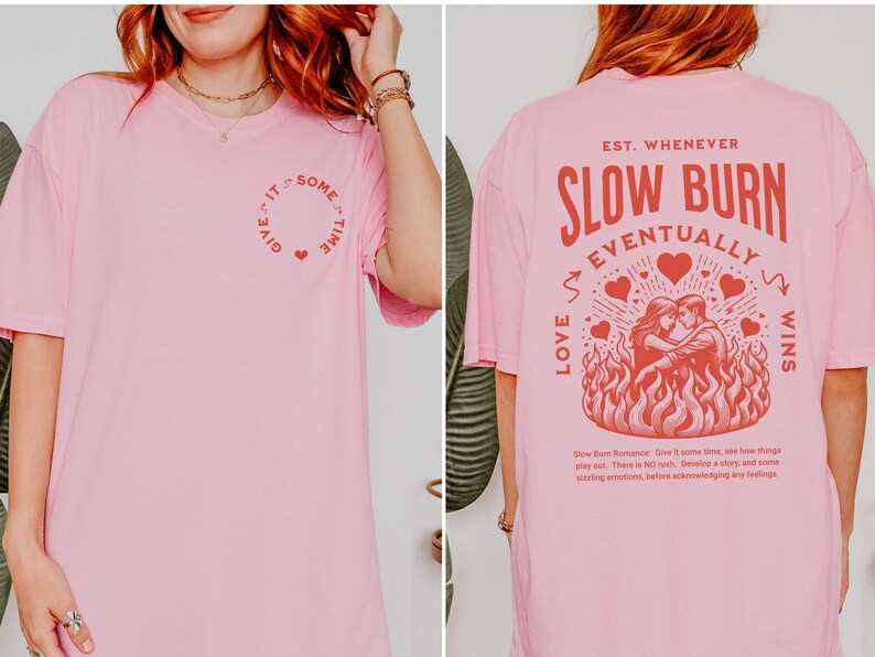 Op de afbeelding: Zachtroze T-shirt met rode tekst en afbeeldingen. De voorkant heeft een cirkelvormig ontwerp met de tekst "Give it some time" en een hart. De achterkant toont "Slow Burn Eventually Love Wins" met een stel in vlammen.