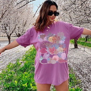 May include: A purple t-shirt featuring a colourful coral reef print. The shirt has short sleeves and a relaxed fit.