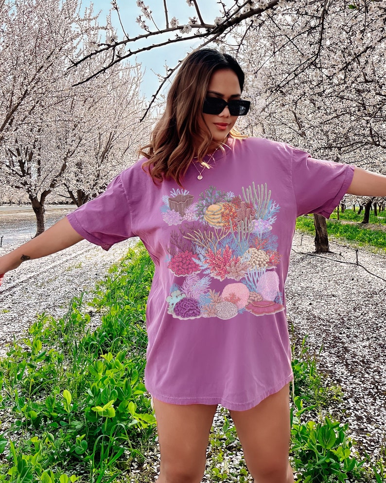 May include: A purple t-shirt featuring a colourful coral reef print. The shirt has short sleeves and a relaxed fit.