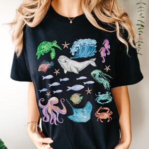 May include: Black t-shirt featuring a colourful ocean-themed graphic print. The design includes a variety of sea creatures, such as a whale, sea turtle, octopus, starfish, and fish.
