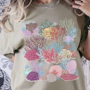 May include: A light green t-shirt featuring a colourful coral reef design. The design includes various types of coral, such as brain coral, staghorn coral, and soft coral. The coral is depicted in shades of pink, red, purple, blue, and green.