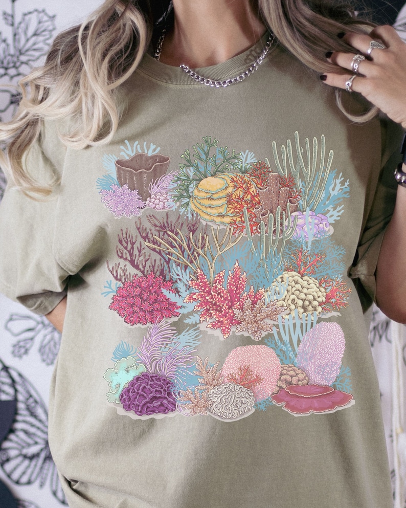May include: A light green t-shirt featuring a colourful coral reef design. The design includes various types of coral, such as brain coral, staghorn coral, and soft coral. The coral is depicted in shades of pink, red, purple, blue, and green.