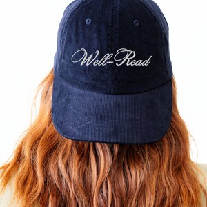 May include: A navy blue corduroy baseball cap with the embroidered text "Well-Read" in white script. The cap has a curved peak and a structured crown. The image shows the back of a person with long, wavy auburn hair.