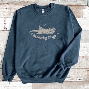 Puede incluir: Una sudadera azul oscuro con un gr&aacute;fico marr&oacute;n claro de una nutria acostada de espaldas con estrellas arriba. El texto "otter-ly fine" est&aacute; debajo de la nutria.