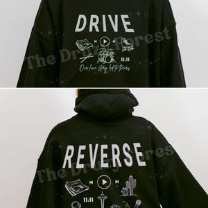 Peut inclure: Un sweat à capuche noir avec le mot "DRIVE" et des icônes telles qu'un journal, un bouton de lecture, une batterie et un patin à roulettes. Le texte "Our love story led to theirs" est en dessous. Le dos dit "REVERSE" avec des icônes similaires.