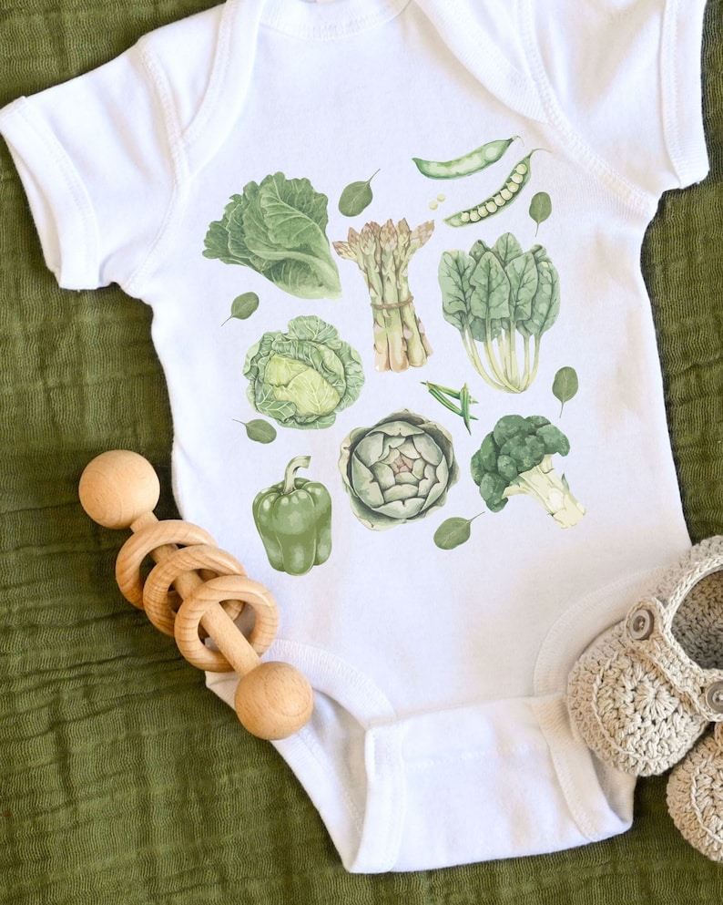 May include: White baby onesie with a green vegetable print. The print includes asparagus, artichoke, broccoli, spinach, cabbage, and bell peppers.