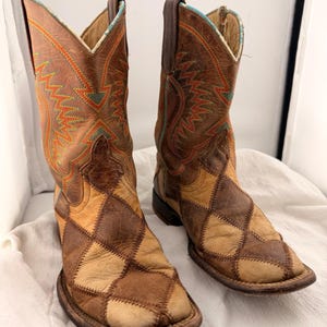 May include: A pair of brown leather cowboy boots. The boots feature a patchwork design on the lower portion and intricate stitching in orange and turquoise on the upper shaft. The boots have a square toe and a stacked heel.