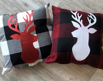 May include: Two decorative throw pillows with a buffalo plaid pattern. One pillow features a red glitter reindeer silhouette, while the other has a white reindeer silhouette.