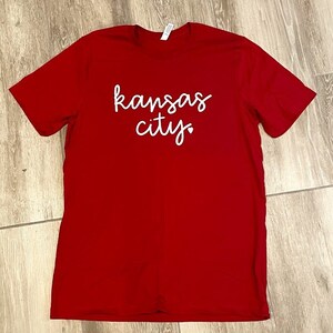 May include: Red t-shirt with white text that says "kansas city" with a small heart after the word "city".