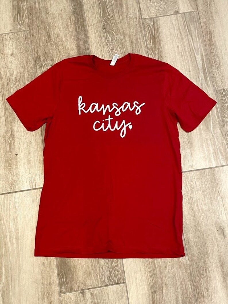 May include: Red t-shirt with white text that says "kansas city" with a small heart after the word "city".