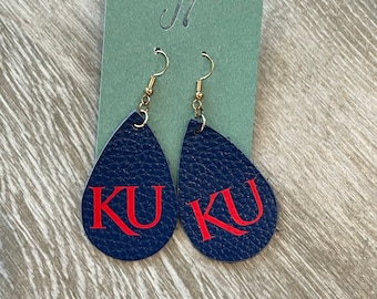 May include: A pair of blue leather earrings with gold hooks. The earrings are shaped like teardrops and have the letters "KU" in red on them.