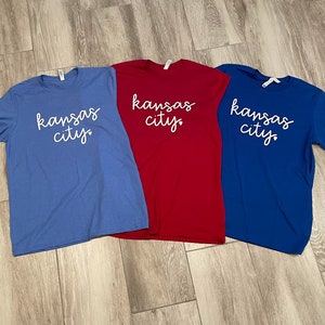 May include: Three t-shirts with the text "kansas city" in white script on a blue, red, and royal blue background.